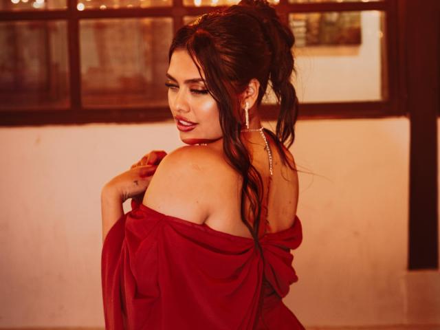 BellaSmithX69 with long dark hair in a ponytail, red off-shoulder dress, pearl necklace, and red lipstick, looking over her shoulder, showing her bare shoulder and slight cleavage.
