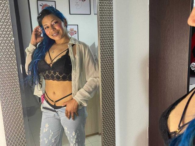 PaulinaColemann with blue hair, smiling, wearing black bra with gold chain, strappy panties, and unbuttoned shirt, reflected in mirror, showing her tits and ass.