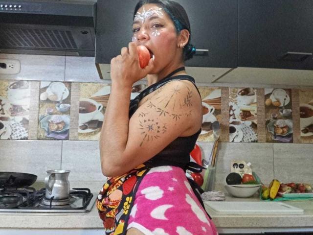 PaulinaColemann, eating an apple, has white face paint and tattoos on her arm. She's wearing a black tank top and pink, white-patterned pants, standing in a kitchen.