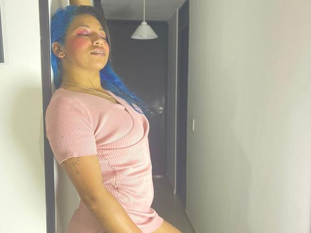 PaulinaColemann with blue hair, pink makeup, and pink ribbed dress, leaning against wall, eyes closed, smiling, showing small tattoo on right arm, in dim hallway.