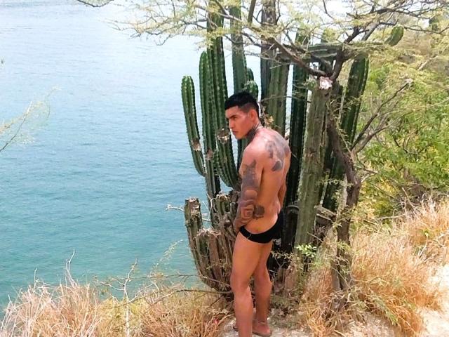 AbrahamStiff with tattoos, black underwear, black cap, and bare chest, standing by a cactus, looking over his shoulder, near a blue ocean, in dry, grassy terrain.