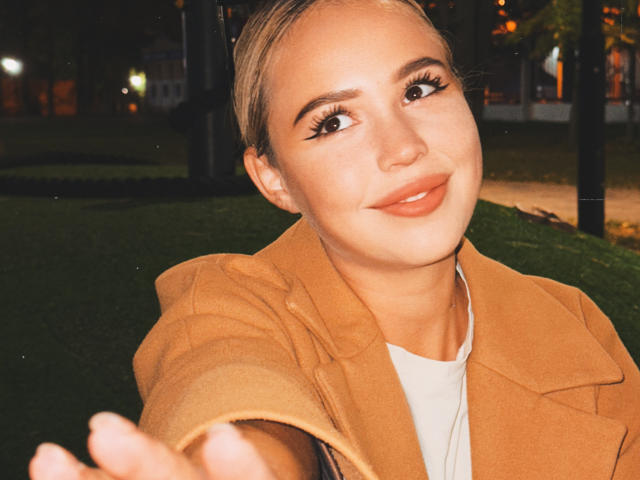 AmyyGlad with blonde hair, brown eyes, and orange lipstick, wearing a tan coat, smiling, reaching out, showing off her tits slightly with her coat open. Nighttime selfie.