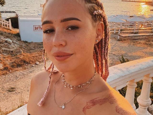 AmyyGlad with pink braided hair, freckles, and a star necklace, showing off her bare shoulders and dragon tattoo, standing by a sunlit waterfront. Her tits are not visible.