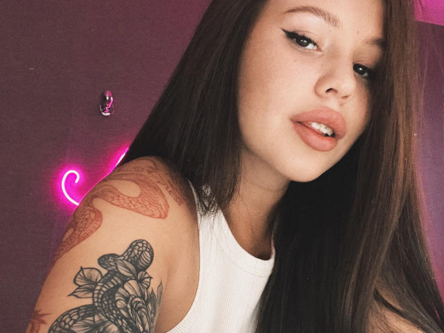 AmyyGlad with long brown hair, white tank top, and pink eyeliner, showing off her tattooed arm with flowers and snake, and slightly parted pink lips.