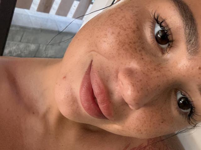AmyyGlad's freckled face with full pink lips, brown eyes, and long eyelashes, looking slightly to the side with a subtle smile. Her bare shoulders are visible.