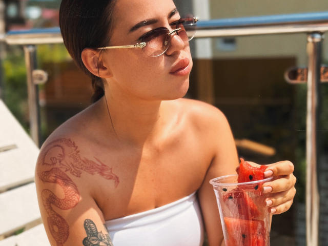 AmyyGlad with a red dragon tattoo on her shoulder, wearing a white strapless top, holding a glass of watermelon, pouting, in sunglasses, on a sunny balcony.