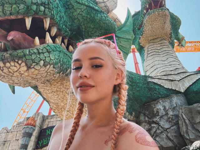 AmyGlad with pink braids, no shirt, showing tattoos, standing in front of a giant green dragon, looking confident, pink sunglasses on head, blue sky background, smiling slightly.