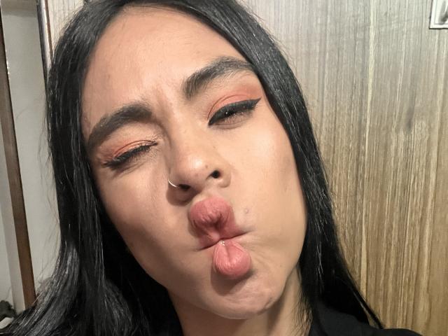 StellaBeckker with black hair, nose ring, and orange eyeshadow, puckering her lips and winking, looking slightly goofy. Her tits are not visible.