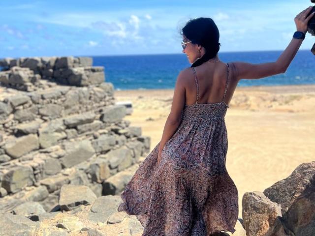 PoisonlLizzy indossa un abito leggero e con motivi, con la schiena rivolta verso la fotocamera, mentre alza il braccio destro, indossa occhiali da sole e si trova accanto a antiche rovine di pietra, con l'oceano sullo sfondo..