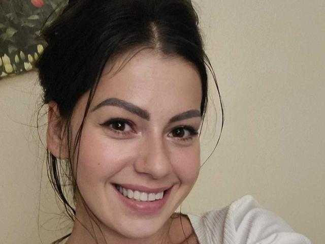 El rostro sonriente de KaylaStorm, con cejas oscuras y gruesas, pestañas largas y labios rosados. Tiene piel clara, cabello oscuro recogido en un moño despeinado, y lleva una blusa blanca..
