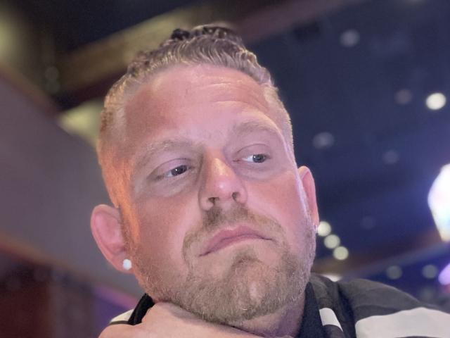 FresherDanny hat ein rundes Gesicht, kurze braune Haare und einen leichten Bart. Er wirkt leicht gelangweilt, wobei sein Kinn auf seiner Hand ruht. Seine Nase ist klein..