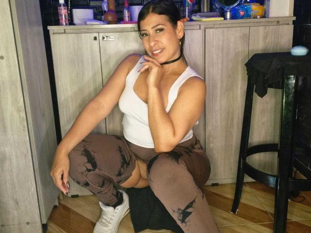 LaurenHills with brown ripped pants, white tank top, and white sneakers, squatting, smiling, black choker, her tits visible, dark hair, wood floor, kitchen background, right hand on chin.