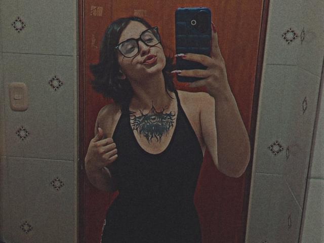 Mirror selfie of MissTat with glasses, black dress, and chest tattoo, holding phone, lips slightly parted, dark hair, red nails, wood door, white tiled walls.