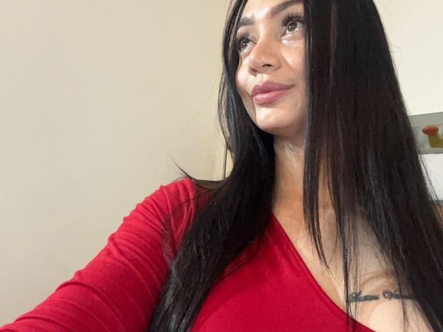SophieLombardo with long black hair, red top, and subtle smile, showing a tattoo on her right chest, with visible makeup and a small tear on her right cheek.