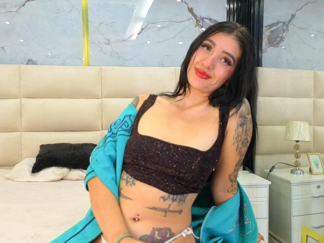 TifanyDiaz with black hair, red lipstick, black sparkly crop top, blue robe, tattoos, and belly button piercing, smiling at the camera, sitting on a bed.