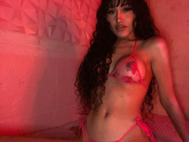 TheaLux with long curly black hair, wearing a pink sheer bikini, showing her small tits and slim ass, under red lighting, looking at the camera with a slightly open mouth.