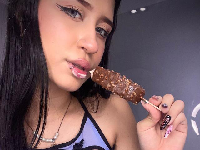 AnnaTorress, con un helado en la boca cubierto de chocolate y nueces, sosteniéndolo con su mano derecha, mostrando su manicura negra y rosa, vestida con una blusa azul y negra, con un piercing en la nariz..