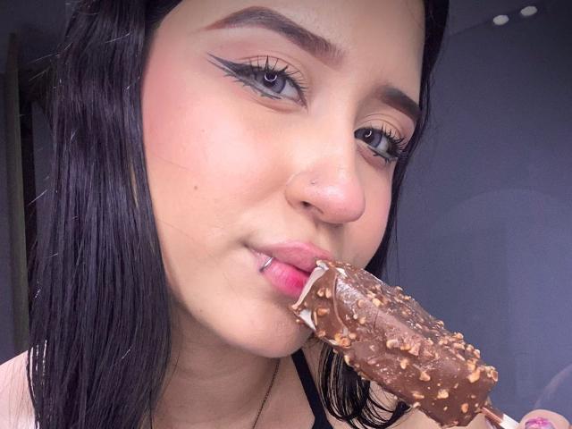 AnnaTorress, con cabello oscuro, delineador de ojos y lápiz labial rosa, comiendo un helado cubierto de chocolate y adornado con nueces, mostrando su lengua rosa y un pequeño piercing en la nariz..