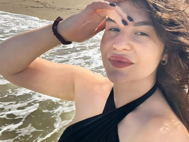 AurikaBriz, with dark brown hair, black nails, and a black halter top, stands by the ocean, touching her head, showing her ass in the sunlight.