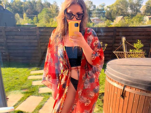 AurikaBriz with wavy brown hair, sunglasses, red floral kimono, black bikini, and phone, showing her bare pussy in a sunny backyard.