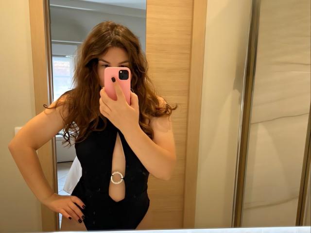 AurikaBriz with wavy brown hair, wearing a black cutout one-piece, holding a pink phone, showing a silver ring in the chest area, standing in a bathroom.