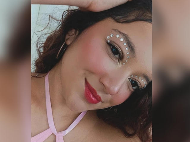 NicoleMartins has curly brown hair, pink makeup with pearl-like dots, red lipstick, and a pink strappy top. She's smiling with hoop earrings, her hand on her head.