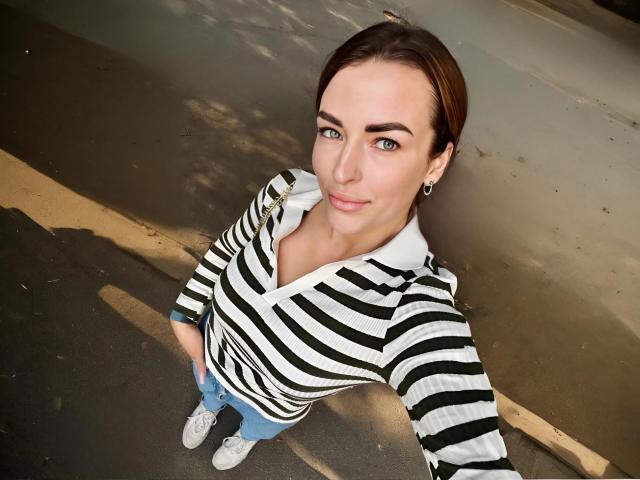 MandiBrooks with striped shirt, blue jeans, white sneakers, and brown hair, looking up with a smile, standing on a sidewalk, wearing a black ear ring.