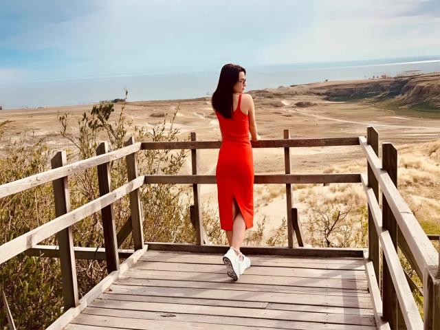 MandiBrooks stands on a wooden bridge, wearing a tight red dress with a slit, showing her ass, and white sneakers, looking at the beach and ocean.