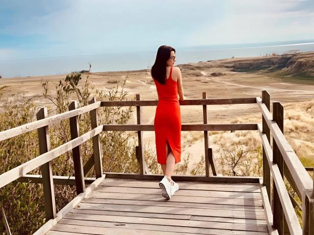 MandiBrooks with ass in a tight red dress, standing on wooden stairs, looking back, wearing white sneakers, dark hair, sunny day, beach and dunes in the background.