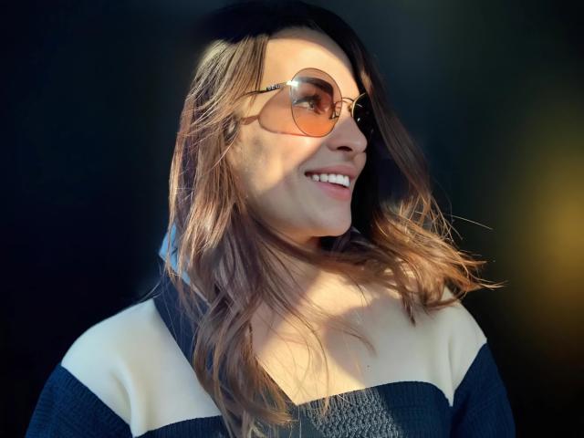 MandiBrooks with brown wavy hair, smiling, wearing round sunglasses and a black-and-white striped sweater, sunlight on her face. Boobs not visible. Her ass is not in the frame.