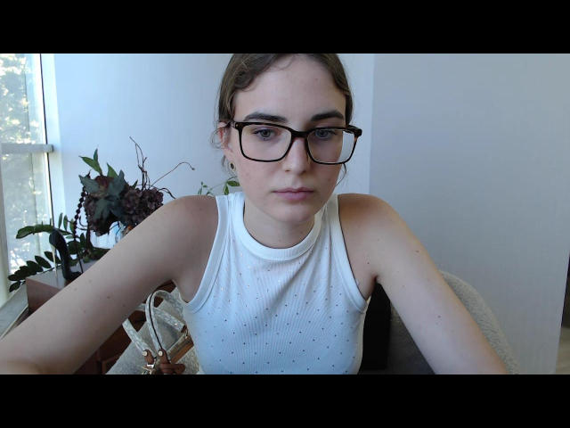 StellaMari with glasses, white dotted sleeveless top, sitting, light skin, brown hair in a bun, looking at camera, arms on table, small plant and window in background.