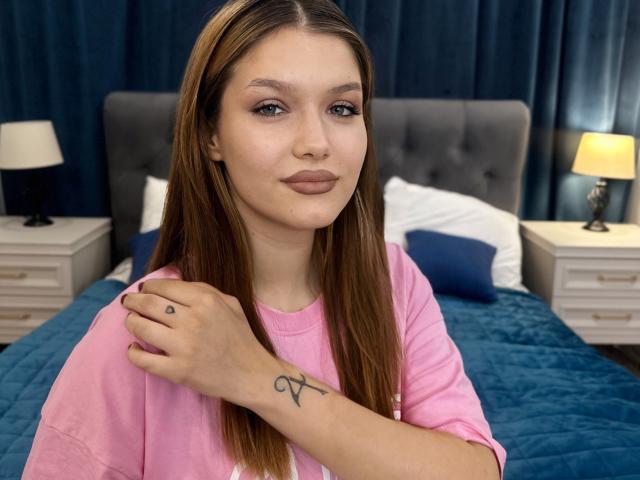 VelvetSkyy with straight brown hair, pink shirt, small tattoo on right wrist, hand on chest, neutral expression, blue bed, white nightstands, yellow lamps, dark blue curtains.