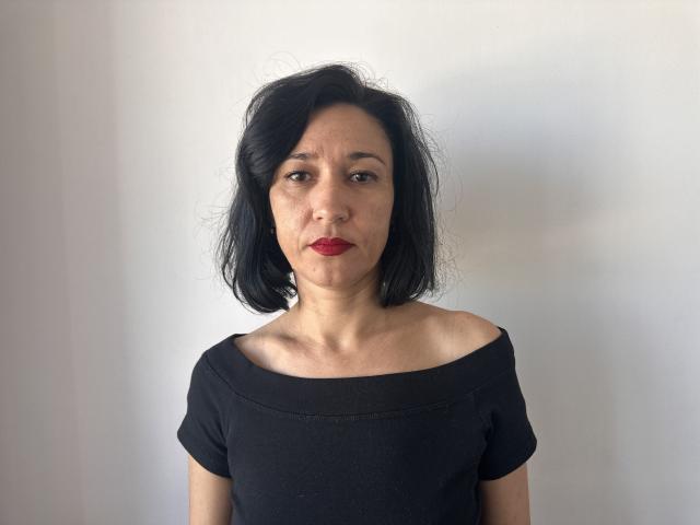 CameronEnns with medium-length black hair, red lipstick, and a serious expression, wearing an off-shoulder black top, showing her tits and ass in a simple, plain background.