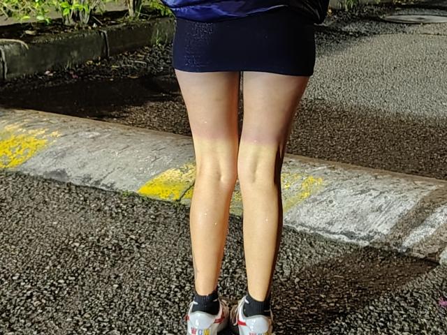 LindaWatsong's legs are standing on a wet, textured road. She's wearing a short, dark blue skirt and white, red, and black sneakers. Her legs have a light, yellowish sheen.