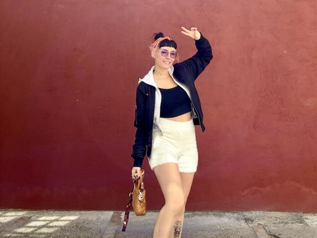 SaraleePoison has pink hair, wearing a black top, white furry shorts, black jacket, red headband, and sunglasses. She's flashing a peace sign, holding a brown bag.