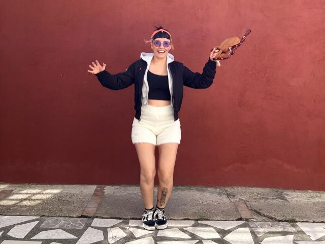 SaraleePoison mit rosa Sonnenbrille, schwarzem Crop-Top, weißen Shorts, schwarzer Jacke, hält ein Taco, lächelt, Arme erhoben, steht auf einem gemusterten Bürgersteig, Tattoo am rechten Oberschenkel..