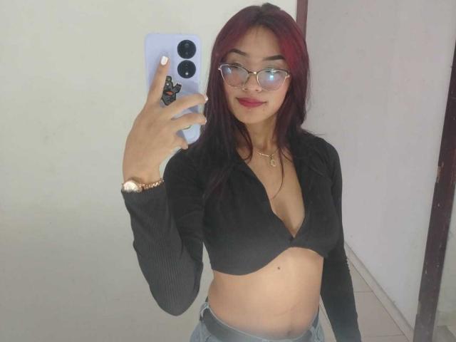 SashaHans with red hair, glasses, and red lipstick, wearing a black ribbed crop top exposing her tits and blue jeans, taking a mirror selfie with a white phone.
