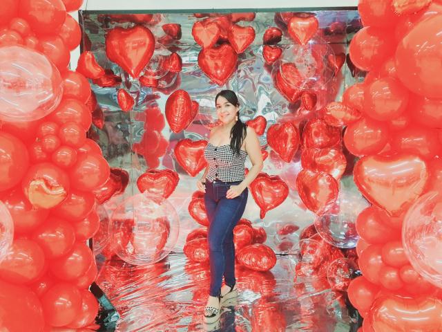 TamaraFoxy standing in a room filled with red heart-shaped balloons, wearing a striped tank top, blue jeans, and beige shoes, smiling with her hands on her hips.