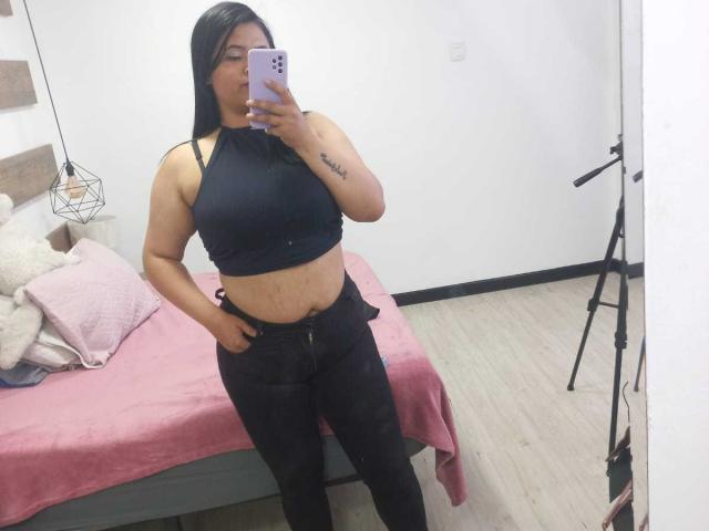 EmilyXGrace with long black hair, wearing a black halter top and black jeans, showing a tattoo on her right arm, standing in a room with a pink bed.