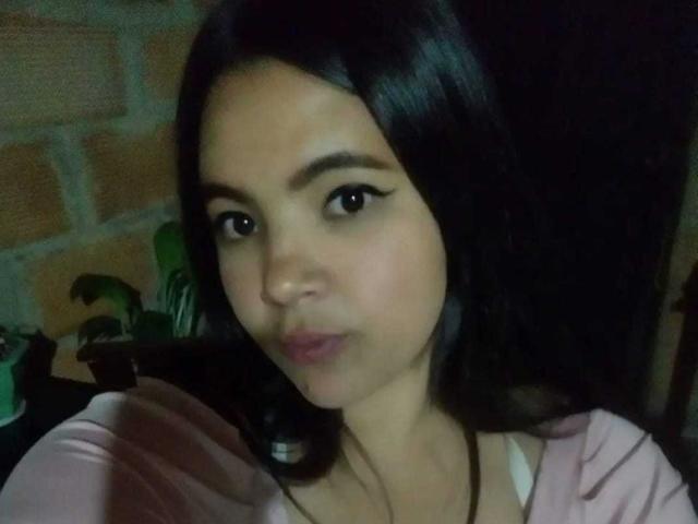 EmilyXGrace has smooth, medium-brown skin, black eyeliner, and black hair. She's pouting slightly with pink lips, wearing a pink top, and looking at the camera.