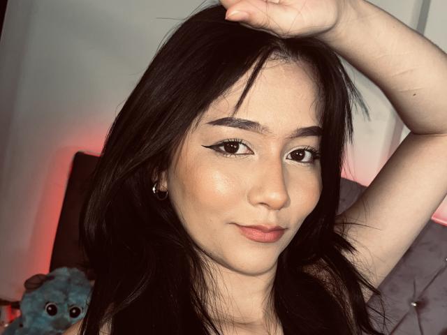 DeisyWest with dark hair, raised arm, and sharp winged eyeliner, showing off her smooth, glowing face and pinkish lips, looking cute and confident. Boobs are not visible.