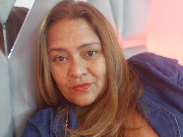 MonzerratBoyer with medium brown hair, red lipstick, and a mole on her upper lip, wearing a blue denim shirt, looking slightly to the side with a soft smile.
