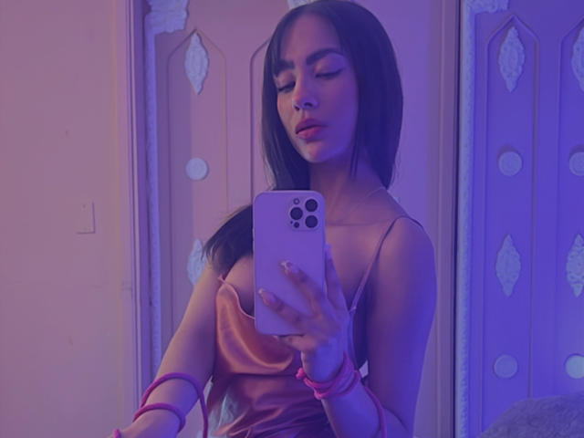 AitanaJohns with dark hair, pink dress, and pink bracelets, holding a pink phone, taking a mirror selfie, eyes closed, showing her tits and nose ring.