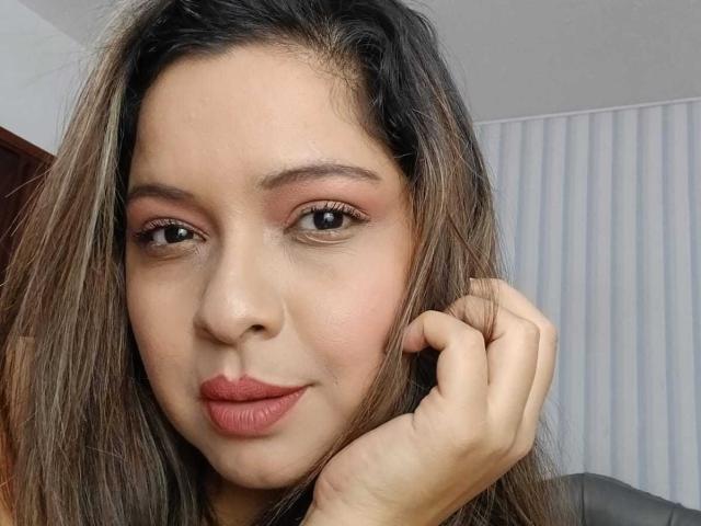 IdyllaJhones has light brown skin, brown eyes, and pink lipstick. She's touching her hair with one hand, showing off her slight smile and natural, wavy brown hair.