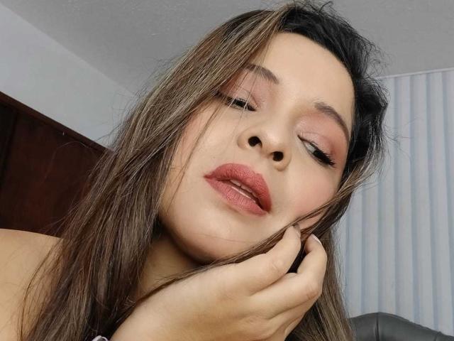 Idylla Jhones, con gli occhi chiusi, capelli castani, ombretto rosa, rossetto rosso, una mano che tocca la guancia, espressione delicata, pelle chiara, leggero sorriso, trucco leggero, casual, interni, rilassata..