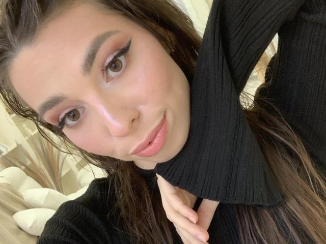 ChristieStorm with pouty pink lips, brown eyes, and winged eyeliner, wearing a black ribbed turtleneck, touching her neck. She has wet brown hair and a cute, sexy face.