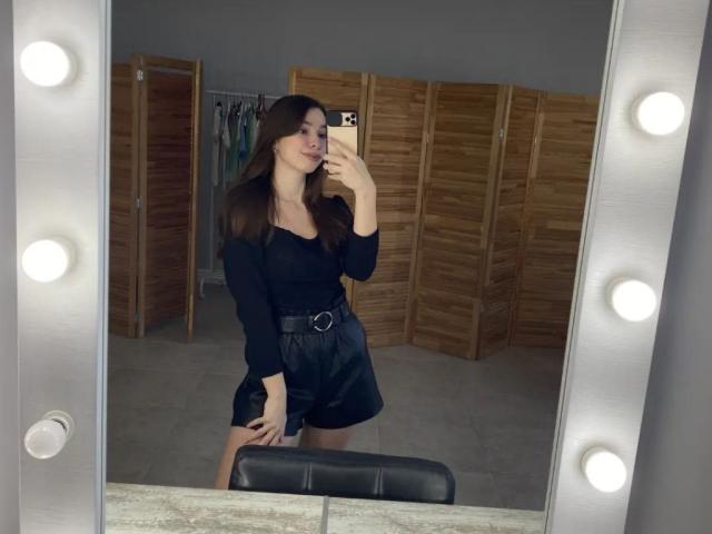 ChristieStorm with long brown hair, black long-sleeve top, black high-waisted shorts, belt, holding phone, mirror selfie, wooden room divider, light bulbs in mirror frame.