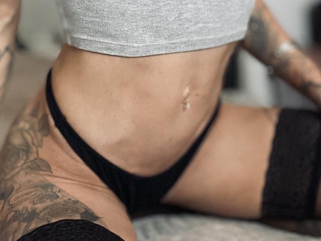 DaneeSoHot's slim, tattooed waist in black panties and gray top, with black lace thigh-highs, showing a belly button piercing.