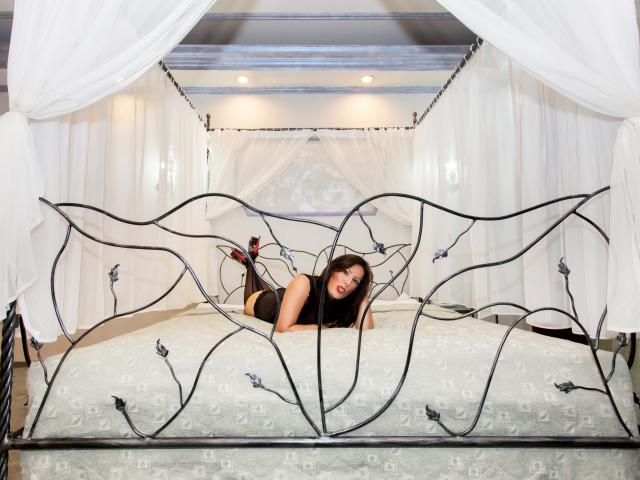 BadAngel with black hair, red lipstick, lying on white bed with ornate black metal headboard, wearing black lingerie, legs up, heels on, looking at camera, sexy, bedroom.
