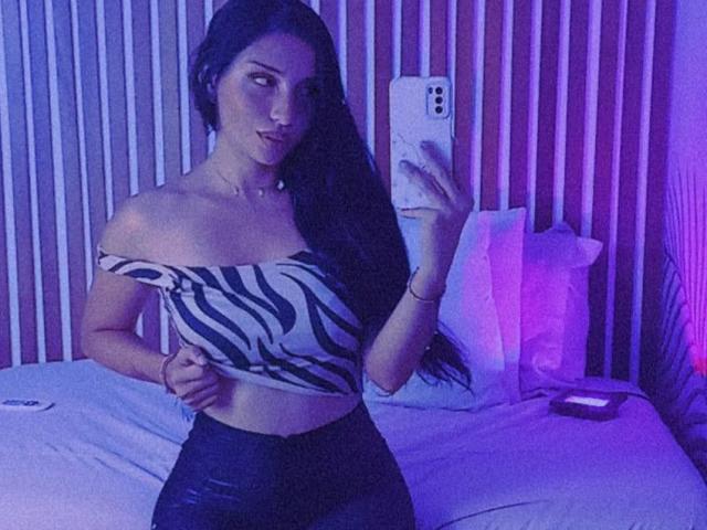 Kate Owens mit langen, dunklen Haaren, einem schwarzen Top mit Zickenzackmuster und freier Schulter, schwarzer Hose, hält ein Telefon, liegt im Bett, violette Beleuchtung, Spiegelbild, sichtiges Gesäß, schlank, attraktiv..